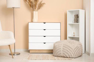 Oturma odasındaki rafların yanında beyaz çekmeceler ve yumuşak puf.