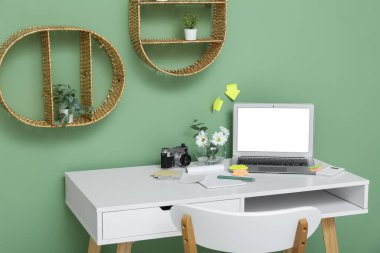 Yeşil duvarın yanında dizüstü bilgisayarı, çiçekleri, kırtasiyesi ve fotoğraf makinesi olan modern ofis işyeri