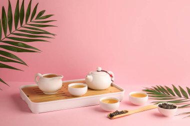 Çaydanlık tepsisi, çay fincanları, kuru yapraklar ve pembe arka planda tropik yapraklar.