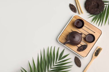 Çaydanlıklı ahşap tepsi, fincanlar, kuru sıkılmış çay ve gri arka planda tropik yapraklar, üst manzara.