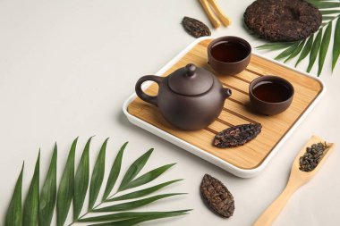 Çaydanlıklı ahşap tepsi, fincanlar, kuru sıkılmış çay ve gri arka planda tropikal yapraklar.