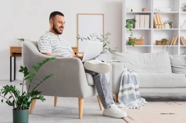 Genç adam evdeki koltukta internette arama yapmak için laptop kullanıyor.