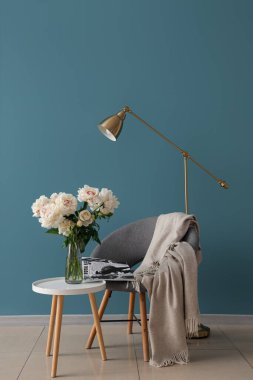 Kahve sehpası, sandalye ve yer lambası olan beyaz şakayık vazosu mavi duvarın yanında.