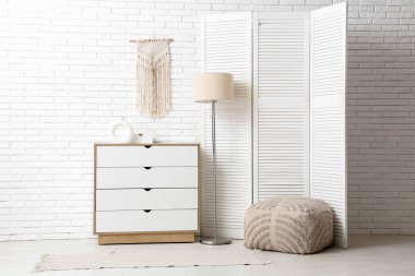 Çekmeceler, katlanır ekran, puf ve zemin lambası, beyaz tuğla duvarın yanında.