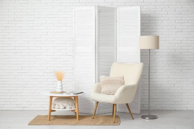 Modern odanın içinde katlanır ekran, koltuk, zemin lambası ve beyaz tuğla duvarın yanındaki masa.