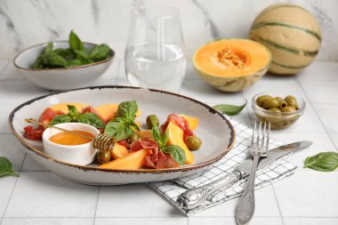 Lezzetli prosciutto, fesleğen yaprağı ve beyaz fayans masasında bal dolu taze kavun parçaları.