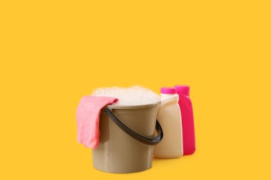 Sarı arka planda köpüklü plastik kova, bez ve şişe deterjan.