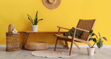 Tahta bank, koltuk, hasır masa ve sarı duvarın yanındaki ev bitkileri.