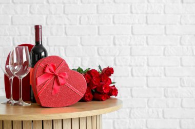 Kalp şeklinde hediye kutusu olan kahve masası, beyaz tuğla duvarın yanında bir buket gül ve şarap. Sevgililer Günü kutlaması