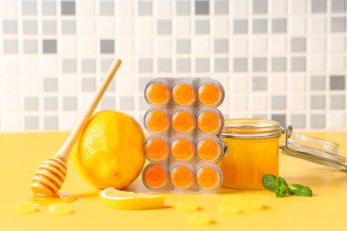 Limonlu öksürük damlaları, bal kavanozu ve nane yaprakları sarı masada gri fayans duvarına karşı.