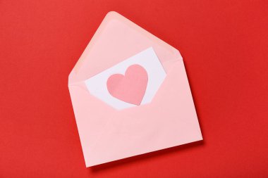 Kırmızı zemin üzerinde pembe zarf, kart ve kağıt kalp olan kompozisyon. Sevgililer Günü kutlaması