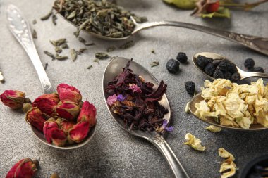 Gri grunge arka planda kaşıklarda farklı türde kuru çay, yakın plan.