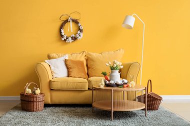 Paskalya için dekore edilmiş oturma odasının içinde sarı kanepe, masa ve lamba var.