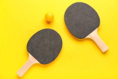 Masa tenisi raketleri ve top sarı arka planda.