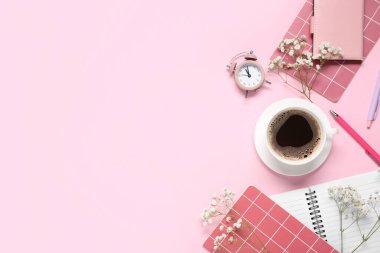 Not defterli bir fincan kahve, çalar saat ve pembe arka planda çingene çiçekleri.