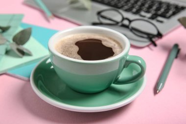 Pembe arka planda dizüstü bilgisayarlı bir fincan kahve, defterler ve gözlük.