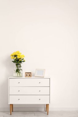 Sarı çiçekli vazo, takvim ve fotoğraf çerçevesi beyaz duvarın yanındaki dolapta. Uluslararası Kadınlar Günü kutlamaları