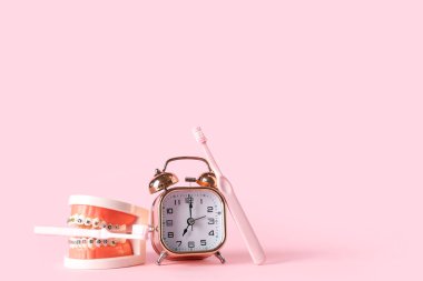 Alarm saati, diş telli çene modeli ve pembe arkaplanda diş fırçası