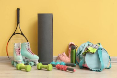 Sarı duvarın yanında farklı spor aletleri, çanta, paten ve spor ayakkabıları var.