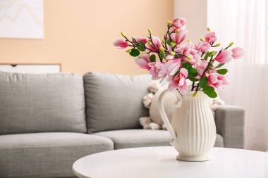 Üzerinde güzel manolya çiçekleri olan vazo şık oturma odasındaki gri kanepenin yanında.