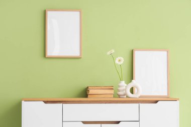 Yeşil duvara karşı klozette papatya çiçekleri, kitaplar ve çerçeveler olan farklı vazolar.