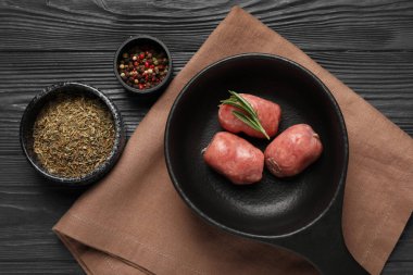 Çiğ sosisli kızartma tavası, biberiye, siyah ahşap arka planda kurutulmuş otlar ve biber taneleri.