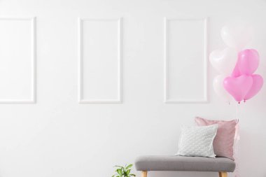 Beyaz duvarın yanında yastıkları ve kalp şeklinde balonları olan gri bir bank. Sevgililer Günü kutlaması