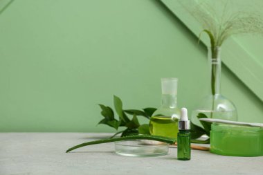 Yeşil duvarın yanındaki gri masada doğal kozmetik ürünlerini hazırlamak için malzemeler ve numunelerle dolu laboratuvar vitrinleri.