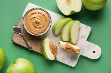 Cutting board with fresh apple slices and bowl of sweet peanut butter on green background