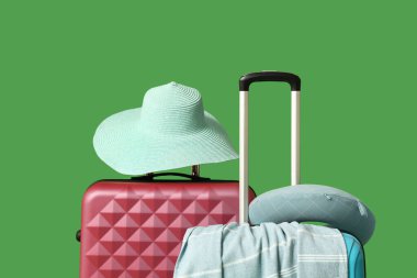 Hasır şapkalı bavullar, yeşil arka planda seyahat yastığı ve havlu.