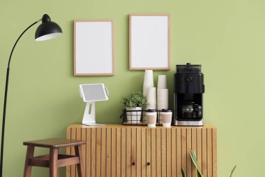 Modern kahve makinesi, tablet bilgisayar ve kağıt bardaklar yeşil duvarın yanındaki dolapta.