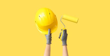 Hands with hardhat and paint roller on yellow background