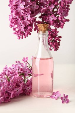 Beyaz duvarın yanındaki ahşap masada güzel leylak çiçekleri olan bir şişe parfüm.