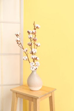 Tahta taburede pamuk filizi vazosu ve sarı duvarın yanında da tuvalet perdesi var.