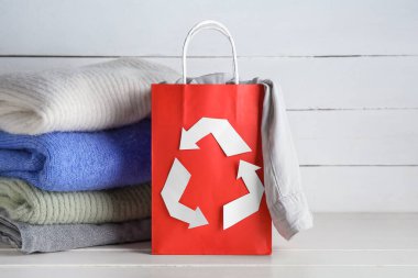 Geri dönüşüm amblemi ve beyaz ahşap arka planda giysileri olan alışveriş torbası.