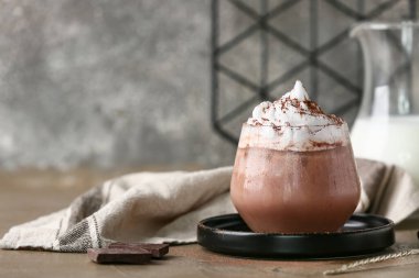 Glass of cocoa drink with whipped cream and chocolate pieces on brown table against grey grunge wall