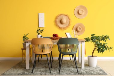 Sarı duvarın yanında sandalyeleri, çiçekleri, suyu ve meyve kasesi olan modern yemek odasının içinde.