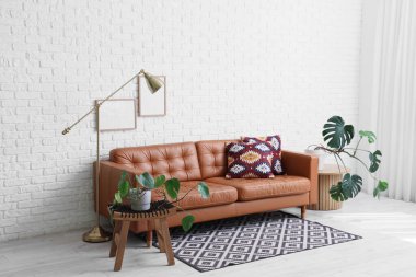 Oturma odasının içinde deri kanepe, lamba ve yeşil Monstera bitkileri kahve masalarında beyaz tuğla duvarın yanında.