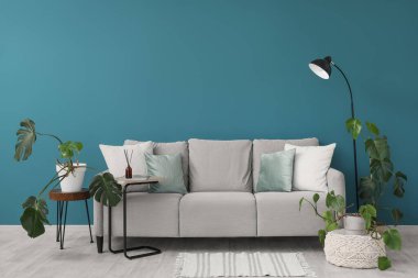 İçinde kanepe, lamba ve yeşil Monstera çiçekleri olan oturma odasının mavi duvarın yanındaki sehpada.