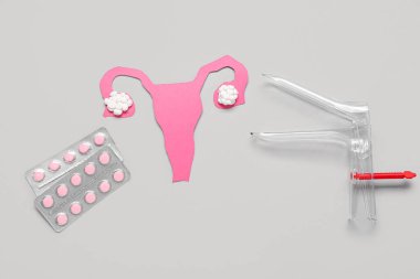 Gri arka planda doğum kontrol hapı ve jinekolojik spekulum içeren kağıt rahim.