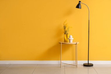 Sarı duvarın yanında çiçekli vazo ve şık standart lamba olan masa.