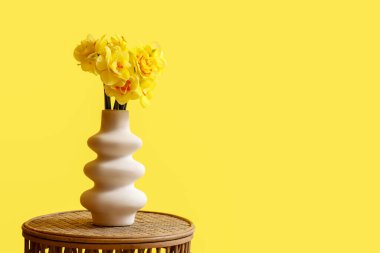 Sarı duvarın yanındaki sehpanın üzerinde bir buket güzel narsisli çiçek olan vazo.