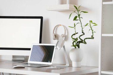 Ofiste beyaz duvarın yanında bilgisayar, dizüstü bilgisayar ve kulaklık bulunan modern işyeri