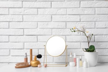 Farklı kozmetik ürünleri, ayna, saç fırçası ve orkide, açık tuğla duvarın yanındaki masada.