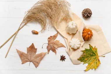 Balkabağı şeklindeki mumlarla sonbahar kompozisyonu, düşmüş yapraklar ve açık ahşap arka planda otlar.