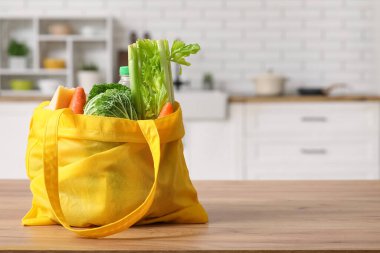Mutfaktaki ahşap masada farklı taze ürünler bulunan sarı alışveriş çantası.