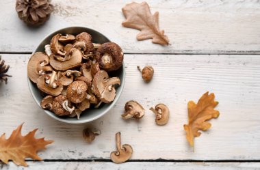 Kurumuş mantarlı kase, sonbahar yaprakları ve bej ahşap arka planda koni.