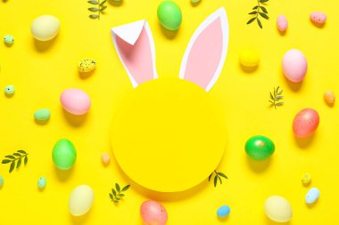 Kağıt tavşan kulaklı boş kart, sarı arka planda Paskalya yumurtaları ve yaprakları.