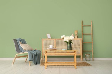 Kahve masasında beyaz şakayık vazosu, sandalye ve şifonyer yeşil duvarın yanında.