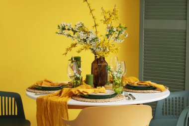 Sarı duvarın yanında çiçek açan dalları olan yemek masası.